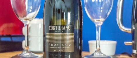 Una botella de Cortigiano Prosecco sobre una mesa de madera, con dos copas de vino y una taza blanca al fondo.
