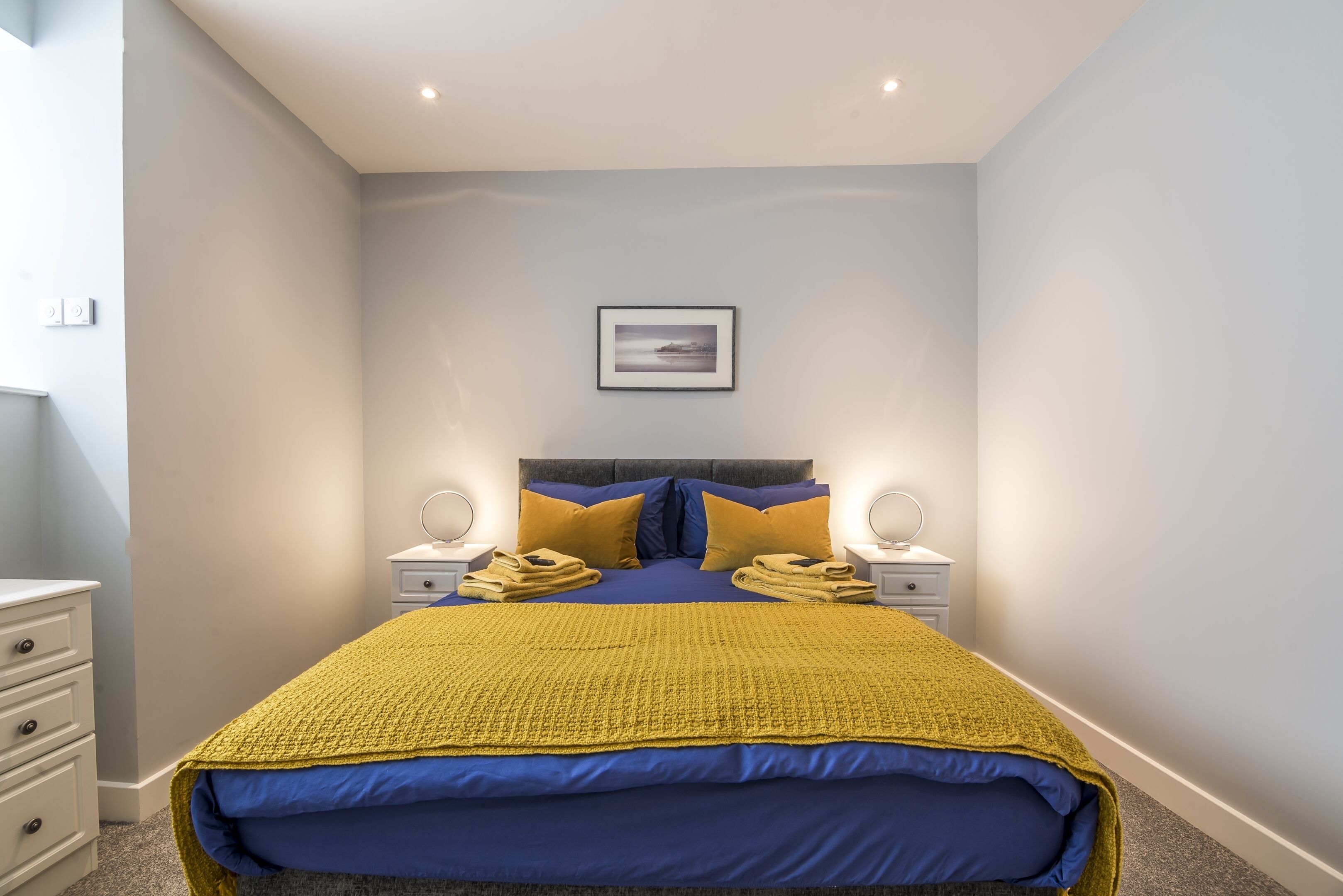 A bedroom with a bed, two nightstands, a headboard, a framed picture on the wall, and a yellow blanket.