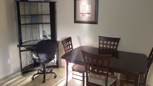 Un comedor con una mesa y sillas de madera oscura, una silla de oficina negra, una estantería y un cuadro en la pared.