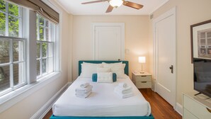 A neatly arranged bedroom with a bed, bedside tables, a television, and a window with curtains. - Oasis Overlooking Forsyth Park w/ Private Balcony & Parking by Lucky Savannah (Savannah)