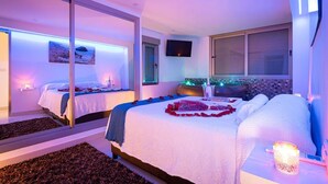 Une chambre d’hôtel moderne dotée d’un grand lit, d’un deuxième lit plus petit, d’une cloison en verre, d’une vue sur la ville et d’un téléviseur.