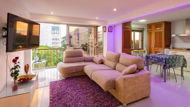Un salon moderne avec un canapé d’angle beige, un téléviseur à écran plat fixé au mur, une table à manger recouverte d’une nappe à motifs floraux et un balcon offrant une vue sur les bâtiments.