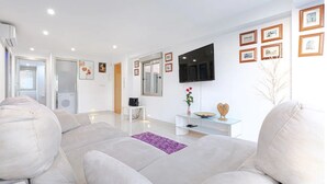 Un salon moderne avec un canapé blanc, une table basse en verre, un téléviseur à écran plat fixé au mur et une petite table d’appoint blanche avec un haut-parleur.