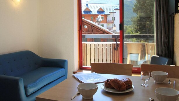 A room with a blue sofa, a wooden table with a plate of pastries, bowls, and a wine glass. A window reveals a mountain view and a building with a red roof. - 20m from ski slopes, 2nd floor, balcony, tv, ski locker, 25m², Serre Chevalier (Briançon)