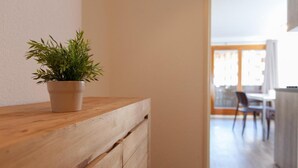 Un comò di legno con una pianta in vaso, un'area pranzo con sedie e tavolo, e vista su un edificio attraverso una finestra.