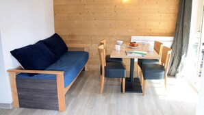 Quarto compacto com piso e parede de madeira, um sofá com almofada, uma mesa de jantar com cadeiras e uma pequena janela.
