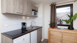 Una cocina compacta equipada con microondas, cafetera y lavavajillas. Se observa una ventana con cortinas y una planta en la encimera.