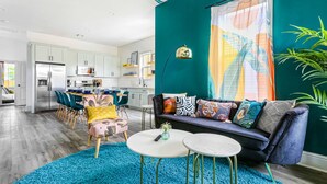 Una cocina moderna con armarios blancos y electrodomésticos de acero inoxidable, una zona de comedor con sillas color turquesa y una sala de estar con sofá azul y cojines estampados.