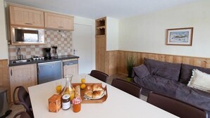 A compact kitchen with wooden cabinets, a microwave, and a coffee maker. A dining table with breakfast items is set in the living area with a couch. - 50m from ski slopes, 3rd floor, balcony, tv, ski locker, 38m², Serre Chevalier (Briançon)