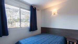 Un lit simple avec une couvre-lit bleue, dans une pièce dotée d’une fenêtre, d’une table de chevet et d’une lampe fixée au mur.