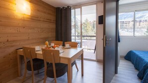 A room with a wooden wall, a dining table with chairs, a bed with a blue blanket, and a balcony with a view of buildings and trees. - 200m from resort center, 2nd floor, balcony, tv, ski locker, 31m², Serre Chevalier (Briançon)