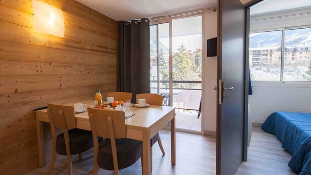 Una habitación con pared de madera, una mesa de comedor con sillas, una cama con cubrecolcha azul y una ventana con vista a edificios y montañas.