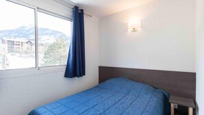 Una cama individual con colcha azul, una ventana con cortina y un aplique de pared.