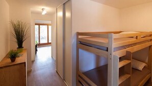 Una cama litera en una habitación con piso de madera, una planta en maceta sobre una mesita auxiliar y una puerta que da a otra habitación.