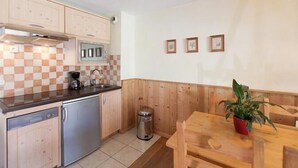 Cocina compacta con gabinetes de madera, refrigerador de acero inoxidable, microondas y un lavamanos. Hay un pequeño cubo de basura y una mesa de comedor de madera con sillas. Una planta en maceta está sobre la mesa.