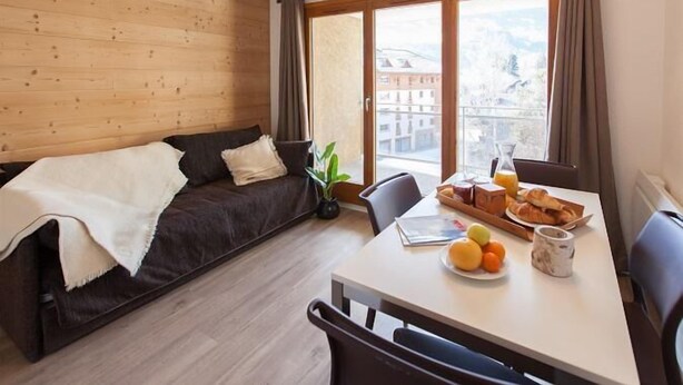 A cozy room with a wooden wall, a dark brown sofa, a dining table with breakfast items, and a large window with a view of buildings and mountains. - 50m from ski slopes, 2nd floor, balcony, tv, ski locker, 30m², Serre Chevalier (Briançon)