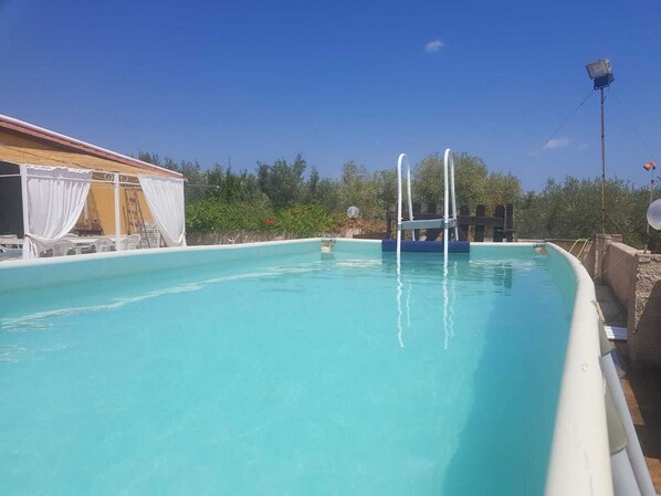 A swimming pool with a clear blue water, surrounded by a white pool deck and a covered area with white curtains.