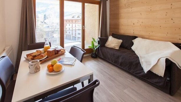 Ein modernes Zimmer mit Esstisch, gedecktem Frühstück, einer Couch und Blick auf ein Gebäude durch ein großes Fenster.
