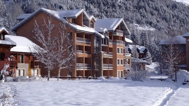 Un immeuble à plusieurs étages doté de balcons, entouré d’arbres couverts de neige et avec une chaîne de montagnes en arrière-plan.