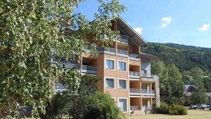 Un immeuble à plusieurs étages, doté de balcons, entouré d’arbres et situé dans un paysage vallonné.