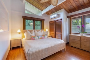 A bedroom with a large bed, wooden floors, a ceiling fan, and a window with a view of greenery.
