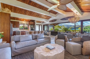 A spacious living area with a sofa, chairs, and a round coffee table. There is a kitchen in the background and a view of the outdoors.