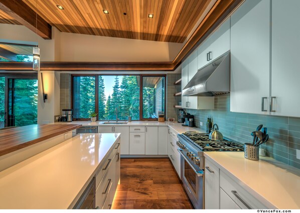 Une cuisine moderne dotée d’un grand îlot central, d’un plafond en bois et de grandes fenêtres donnant sur une forêt.