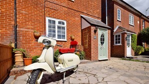 A vintage scooter parked in front of a brick house with a green door and a window with white shutters.