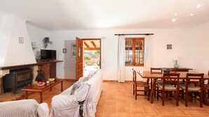 A living room with a fireplace, a television, a dining area with a table and chairs, and a view through an open door. - L´Embat ( Tolo ) Charming 2 bedroom villa (Pollensa)