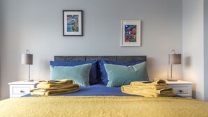 Un lit bien fait, avec une literie bleue et jaune, deux lampes de chevet et deux œuvres d’art encadrées accrochées au mur.