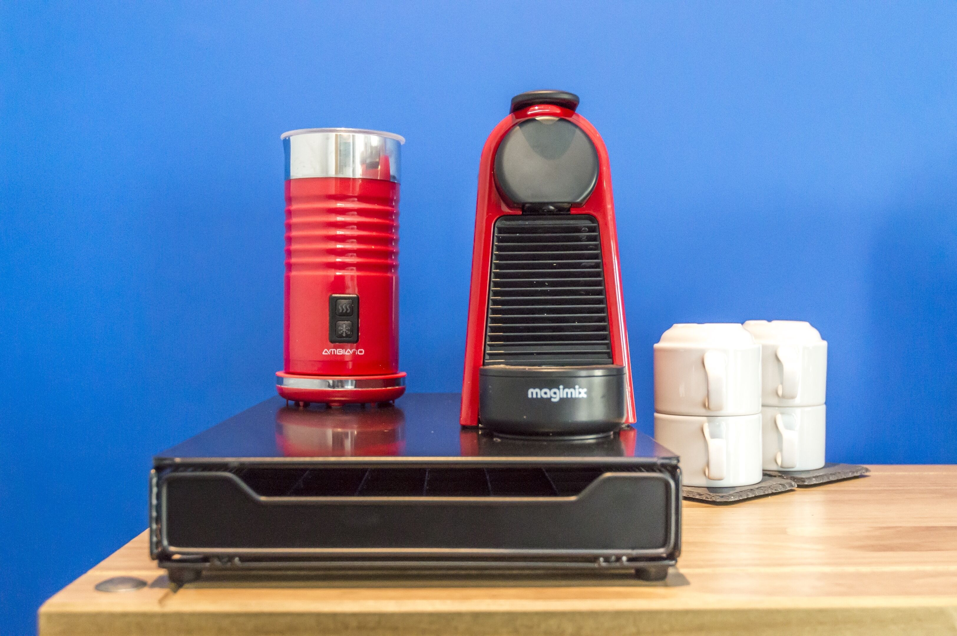 A red and black coffee maker, a stack of white cups, and a black device on a wooden table against a blue background.