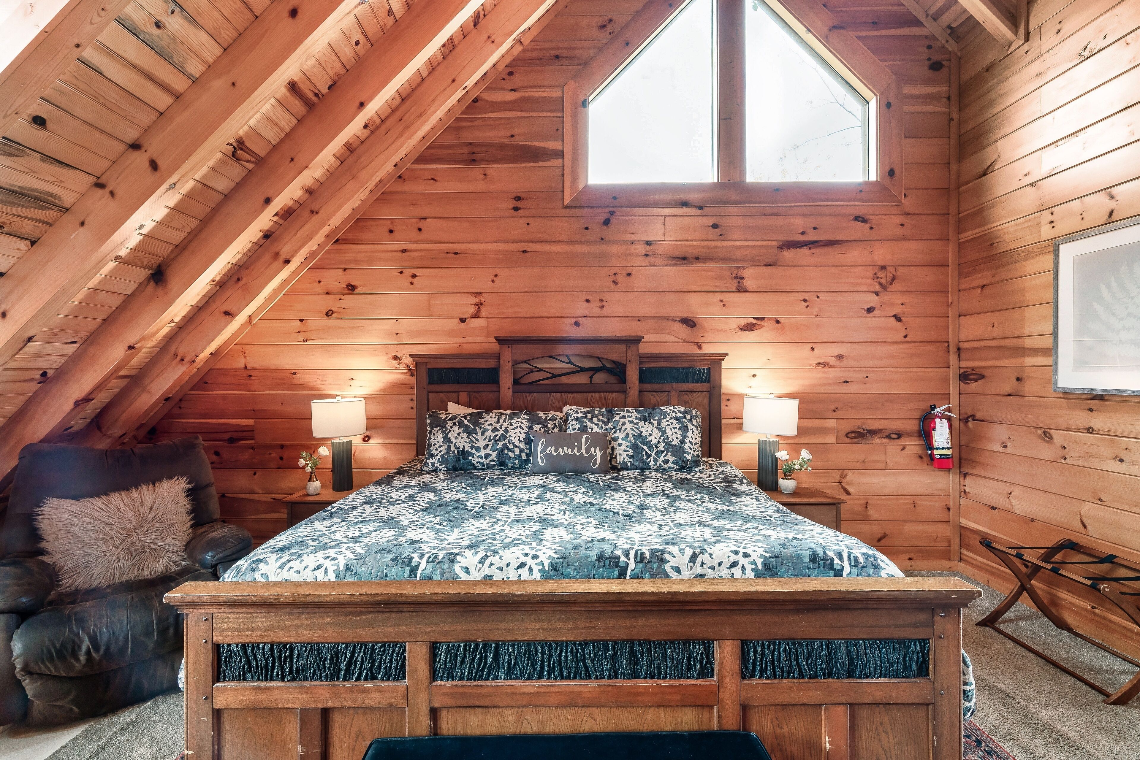 Ein gemütliches Schlafzimmer im Dachgeschoss mit holzgetäfelter Schräge, einem großen Fenster, einem Bett mit gemusterten Betttextilien, einem Nachttisch mit Lampe und einem Ledersessel.