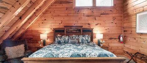 Ein gemütliches Schlafzimmer im Dachgeschoss mit holzgetäfelter Schräge, einem großen Fenster, einem Bett mit gemusterten Betttextilien, einem Nachttisch mit Lampe und einem Ledersessel.
