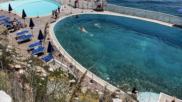 Ein Poolbereich mit klarem, blauem Wasser, umgeben von Liegestühlen und Sonnenschirmen, mit Blick aufs Meer.