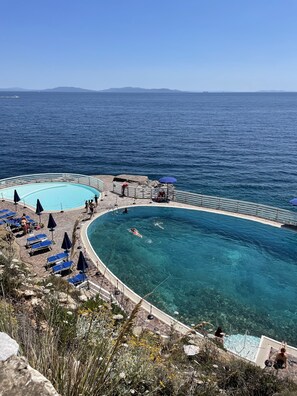 Área de piscina com água azul cristalina, rodeada por espreguiçadeiras e guarda-sóis, com vista para o mar.