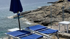 Blick aufs Meer mit felsigem Gelände und ruhiger See. Mehrere blaue Liegestühle mit weißen Gestellen und blauen Kissen, einige mit Sonnenschirmen.