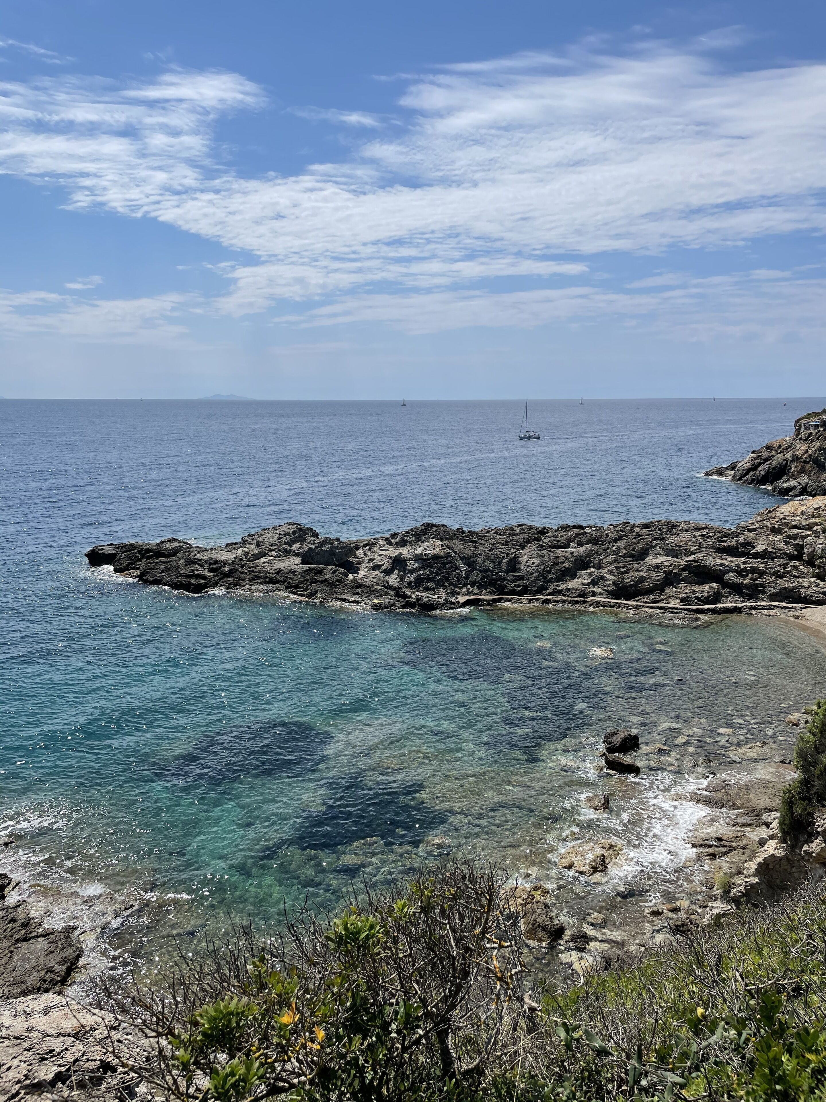 Eine felsige Küste mit klarem, blauem Wasser, ein kleines Segelboot in der Ferne und vereinzelte Vegetation.