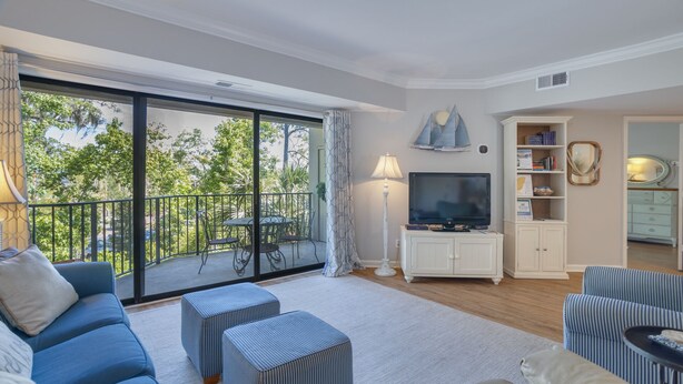 A spacious living room with a balcony, a television, and a bookshelf. - Sunny Coastal Villa| Private Decks, Pool, Tennis | Near Dining & Scenic Grounds (Hilton Head Island)