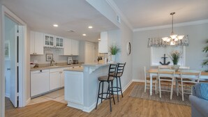 A modern kitchen with white cabinetry, a central island with bar stools, and a dining area with a wooden table and chairs. - Sunny Coastal Villa| Private Decks, Pool, Tennis | Near Dining & Scenic Grounds (Hilton Head Island)