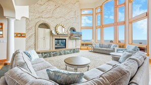 A spacious living room with a large sectional sofa, a fireplace, and a view of the ocean through large windows. - Premier Oceanfront Dog Friendly w/ Pool, Elevator, Hot Tub, Theater, Game Room (Hatteras)