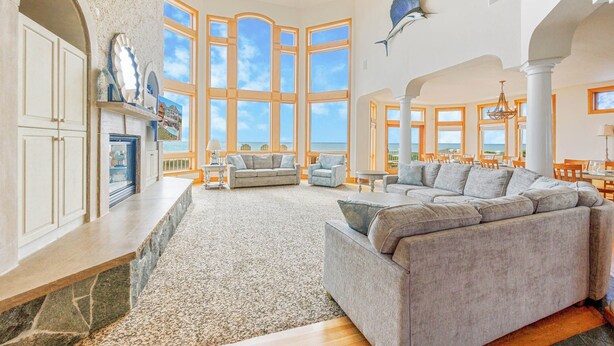 A spacious living area with large windows offering ocean views, a fireplace, and a modern kitchen. - Premier Oceanfront Dog Friendly w/ Pool, Elevator, Hot Tub, Theater, Game Room (Hatteras)