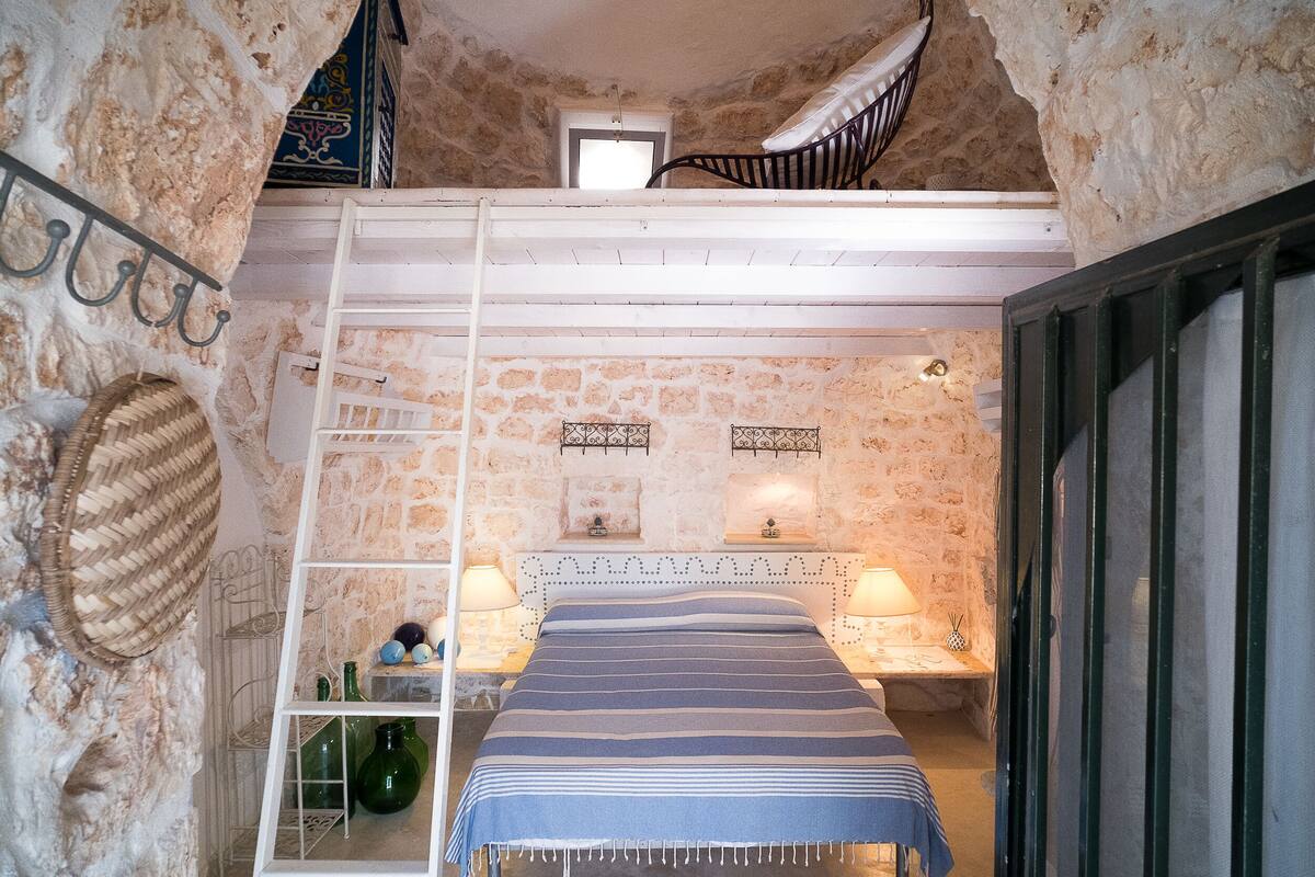 Un dormitorio con pared de piedra, una cama con colcha a rayas, dos lámparas de noche, un ventanal pequeño y una escalera que conduce a un nivel superior.