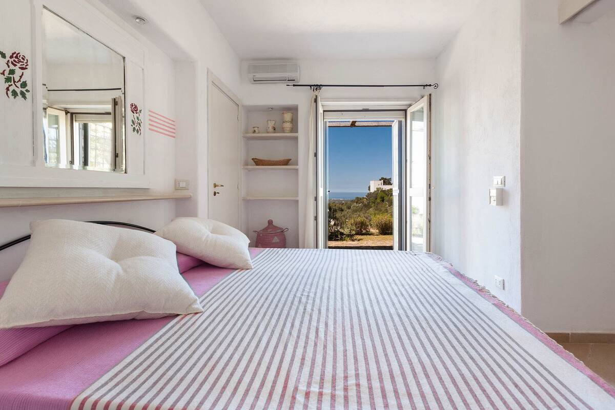 Un dormitorio con cama, almohadas, un aparador y una estantería con objetos decorativos. Se ve una ventana con vistas al exterior.