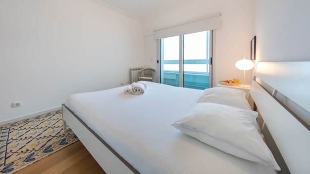 Habitación de hotel con una cama grande, vista al mar a través de una puerta corrediza, una mesita con una lámpara y una silla.