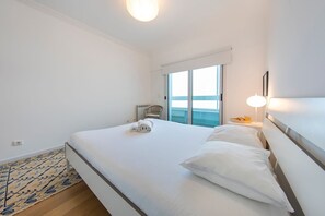 A hotel room with a large bed, a view of the sea through a sliding door, a small table with a lamp, and a chair.