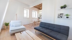 A modern living room with a grey sofa, a white sofa, and a wooden coffee table. There are two windows, a kitchen area, and a dining table with chairs. - Historial Apartment Santos IV (Lisboa)