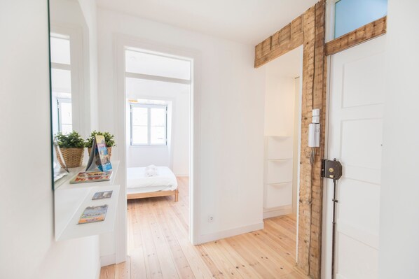 A bright hallway with wooden flooring, a white door, and a bed in the background.