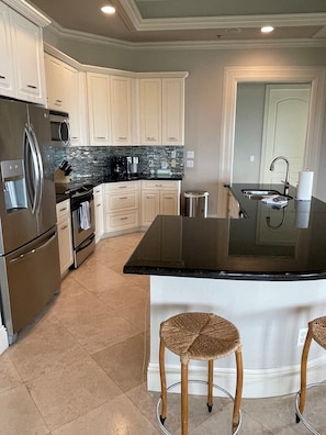 A modern kitchen with white cabinets, stainless steel appliances, a black countertop island, and two wicker stools.