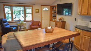 Un coin repas avec une table en bois et quatre chaises, un canapé, un fauteuil et un téléviseur fixé au-dessus d’un comptoir de cuisine.