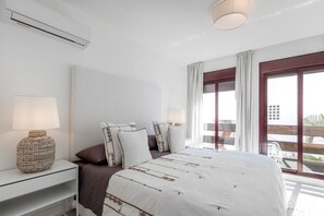 A bedroom with a large bed, a bedside table with a lamp, a window with curtains, and a view of the outside. - La Maestranza | Puerto Banus Townhouse Bbq Pool (Marbella)